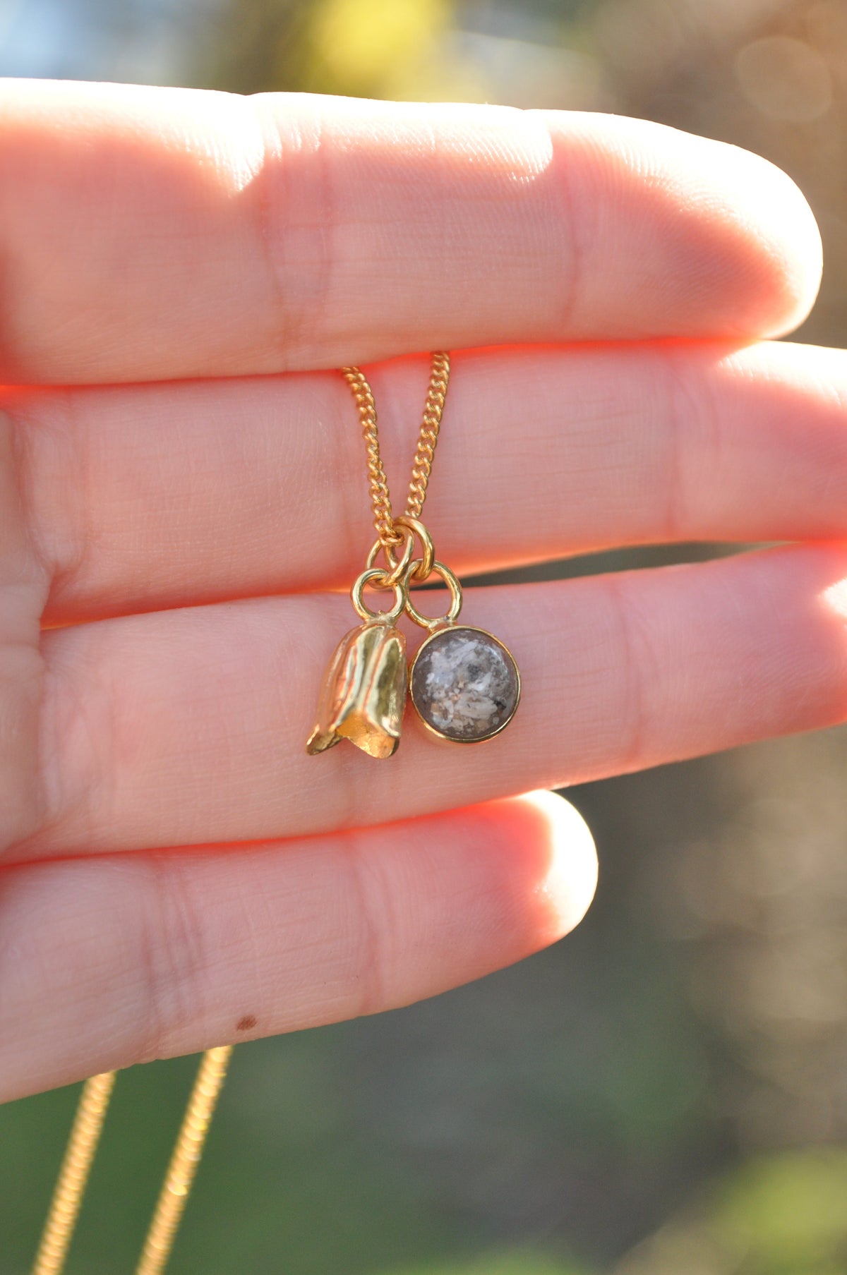 Gold memorial necklace with a delicate floral charm and small round pendant containing ashes, held in a hand in warm sunlight
