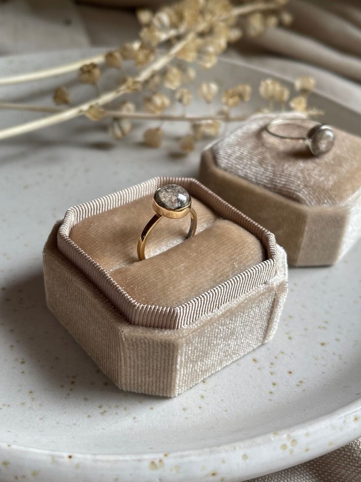 A gold ashes ring displayed in a beige velvet ring box, with a second ring box and a matching ring in the background on a speckled ceramic plate.