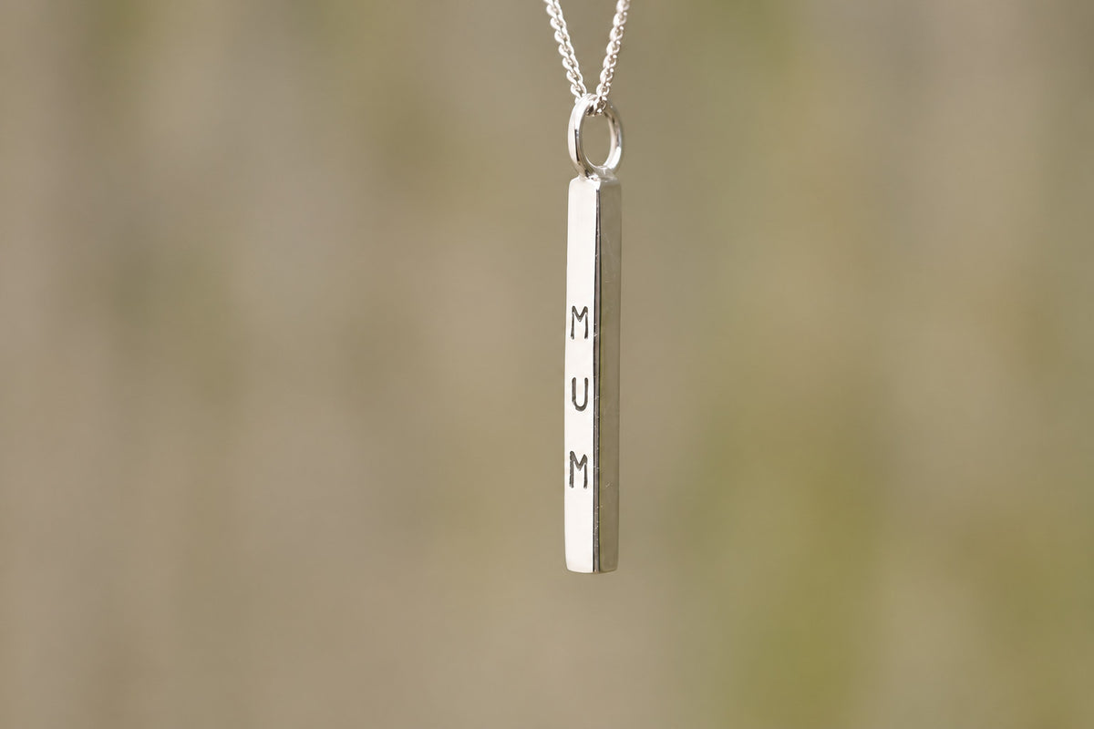 A silver bar pendant necklace engraved with the word 'MUM' vertically, hanging from a delicate silver chain against a soft, blurred beige background.