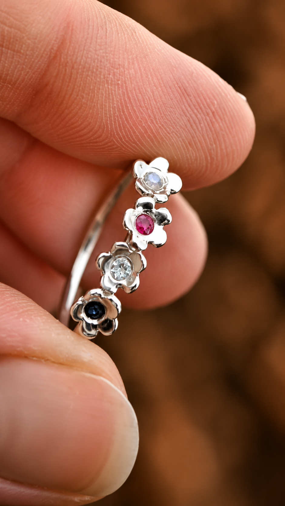 A close-up of a hand holding a silver flower ring with small gemstones in white, pink, clear, and black settings, against a blurred brown background.