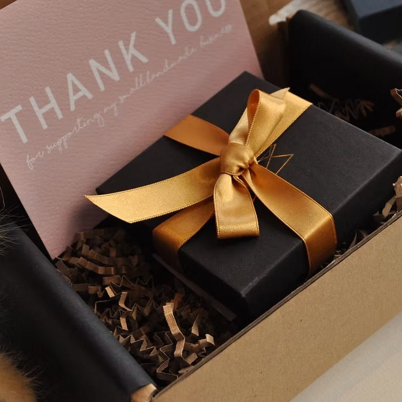A black gift box tied with a shiny gold ribbon and bow, nestled in brown crinkle paper inside a kraft gift box, with a pink thank-you card partially visible.