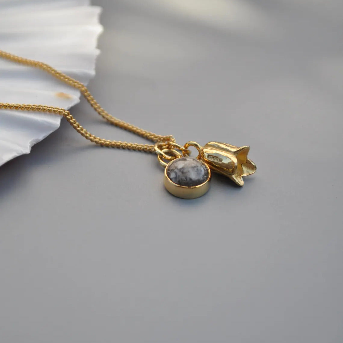 A gold chain necklace with two charms: a round gray memorial ashes pendant and a small textured gold flower charm, displayed on a gray surface with a white decorative leaf in the corner.