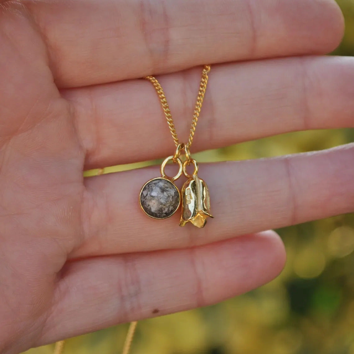 A gold chain necklace with two charms, including a round gray cremation pendant and a textured gold bluebell charm, held in a person's hand against a blurred green background.