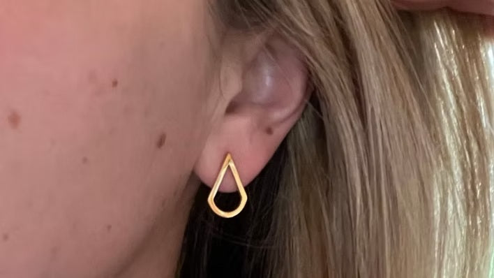 A close-up of a person's ear wearing a small open teardrop-shaped gold hoop earring.