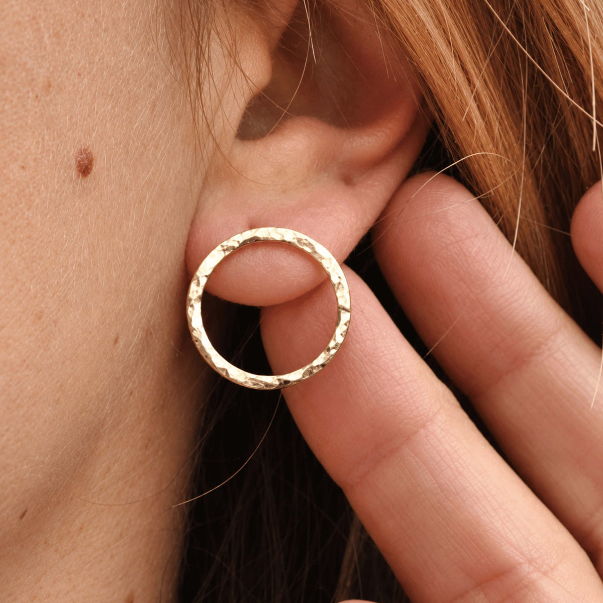 A close-up of a person's ear wearing a textured gold hoop earring, with fingers gently holding the earlobe to display the jewellery.