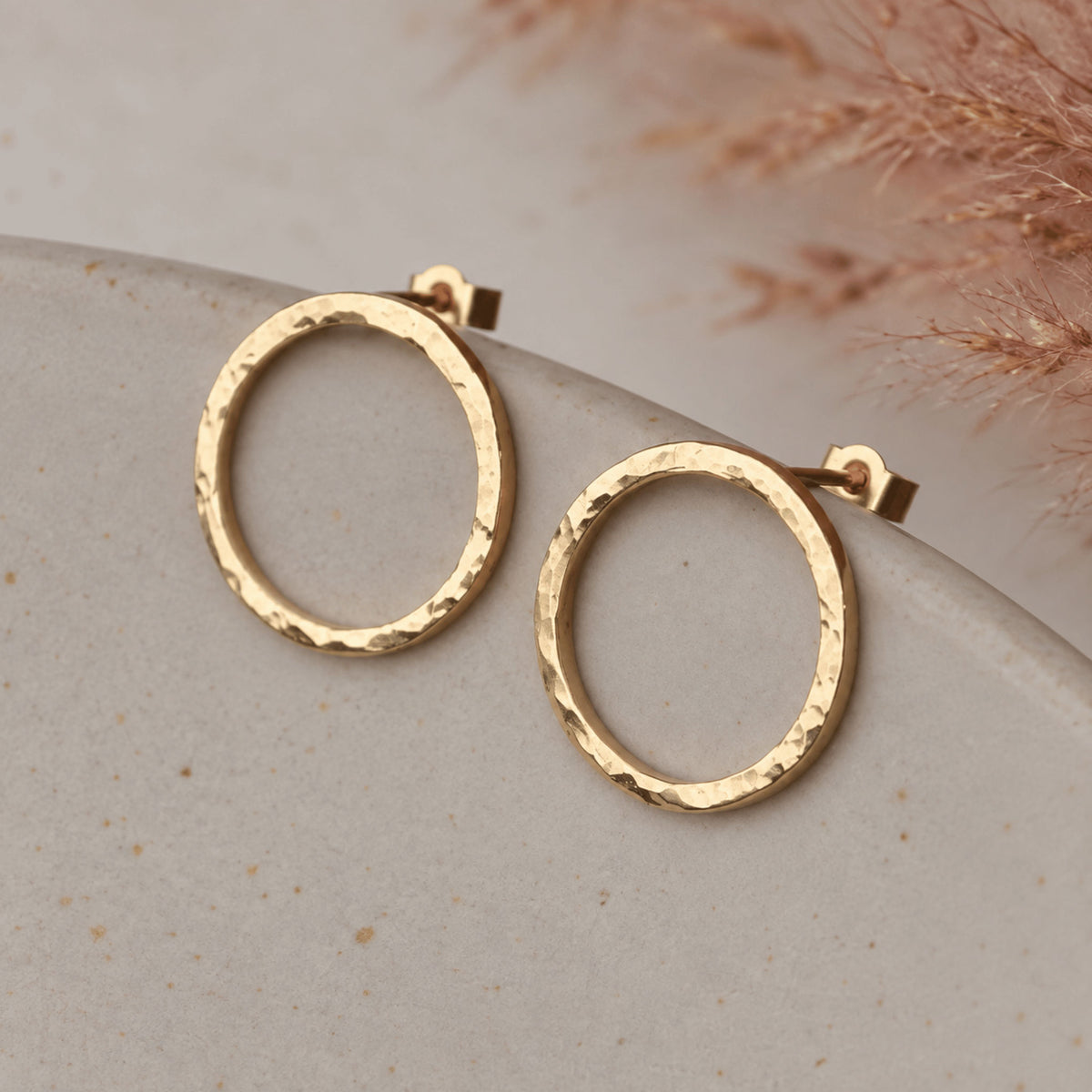 A pair of hammered gold-tone hoop earrings resting on a light beige stone surface with dried pampas grass in the background.