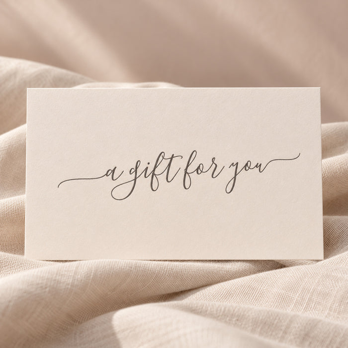A square cream gift card with elegant black cursive script reading 'a gift for you', resting on soft beige fabric.