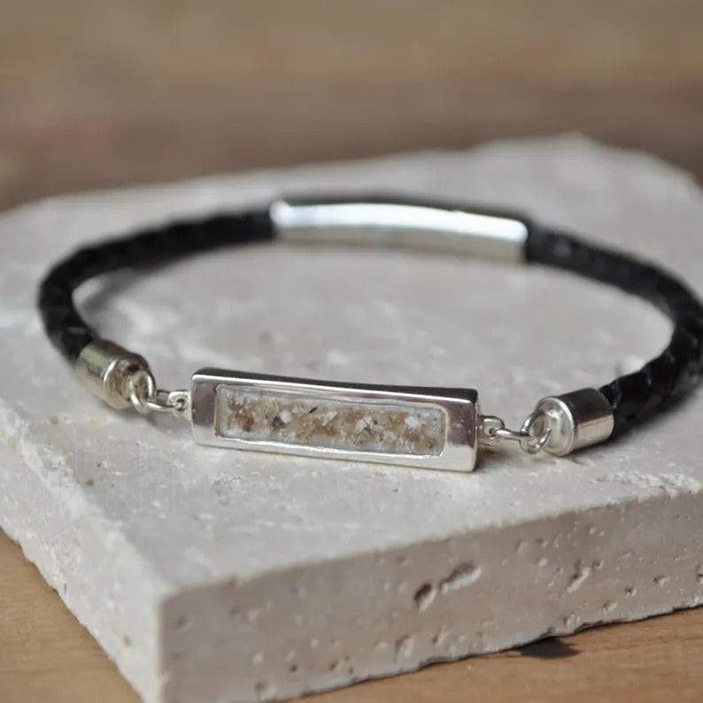 A black braided leather bracelet with a rectangular silver-toned bezel containing a textured beige and brown ashes inlay, displayed on a light stone slab.