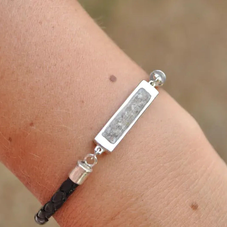 A close-up of a person's wrist wearing a silver-toned rectangular bar bracelet with a bespoke cremation inlay, and a black braided cord detail.
