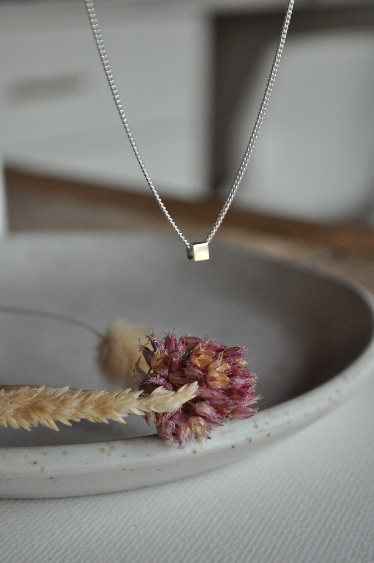 Minimalist silver cube pendant necklace hanging beside dried flowers.
