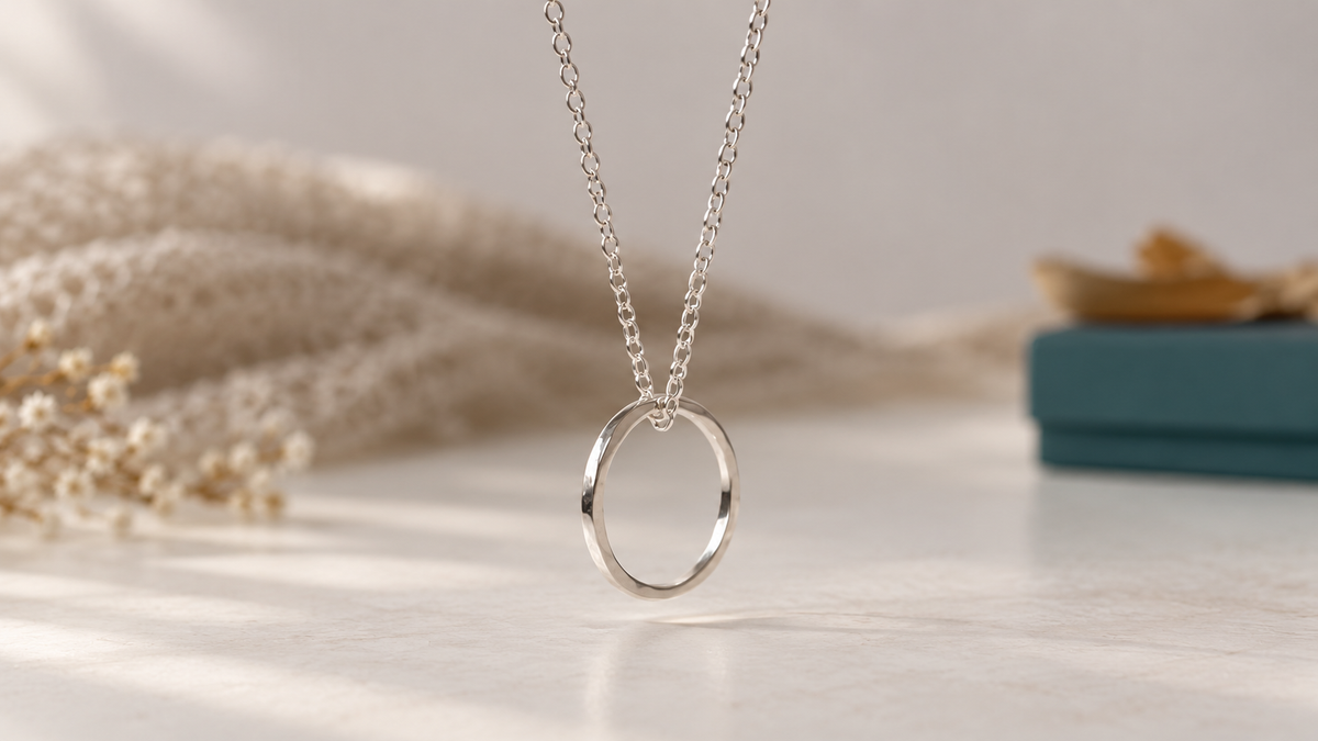 A silver chain necklace with a polished circle pendant, displayed on a light marble surface with soft-focus dried flowers and a teal box in the background.