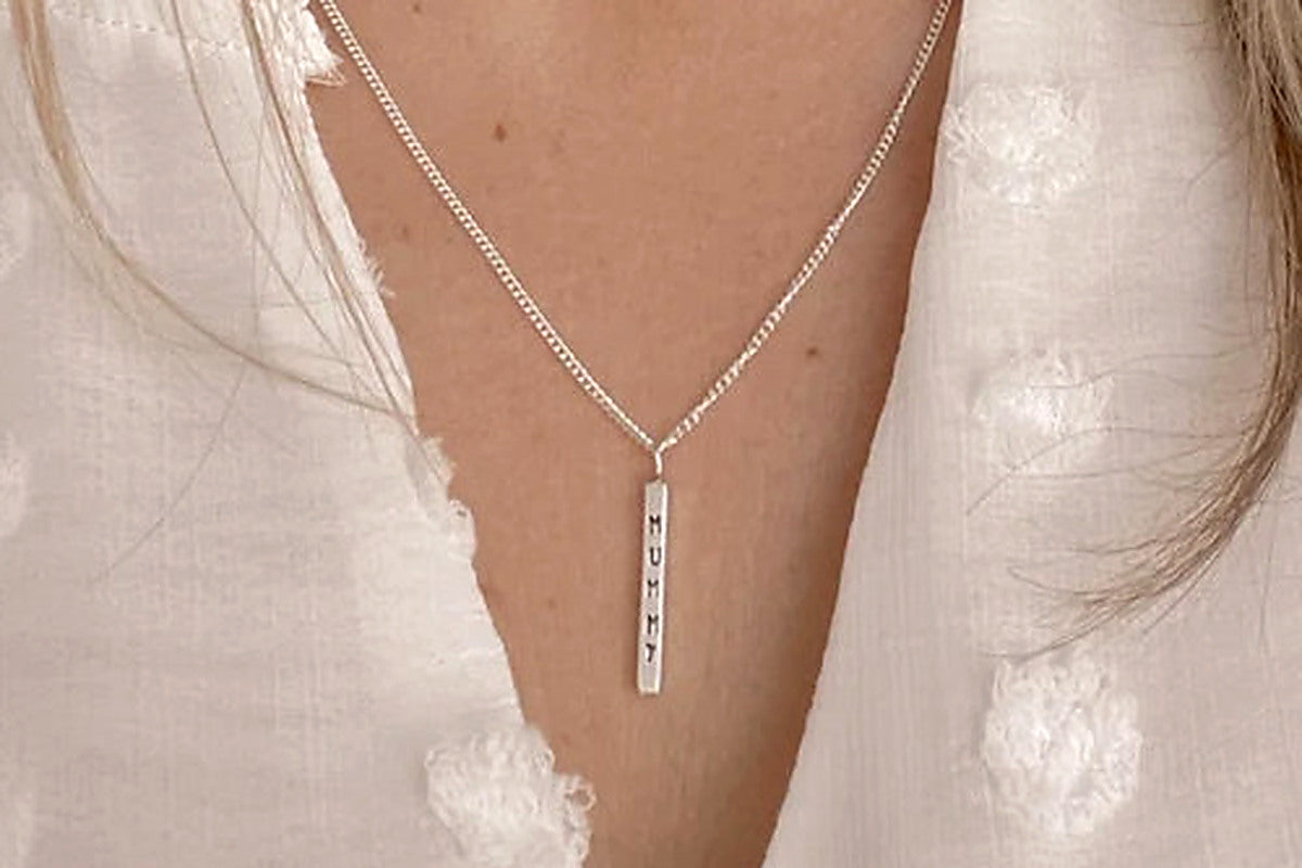 A close-up of a person wearing a delicate silver chain necklace with a vertical bar pendant engraved with 'MUMMY'. The person is wearing a white textured blouse.