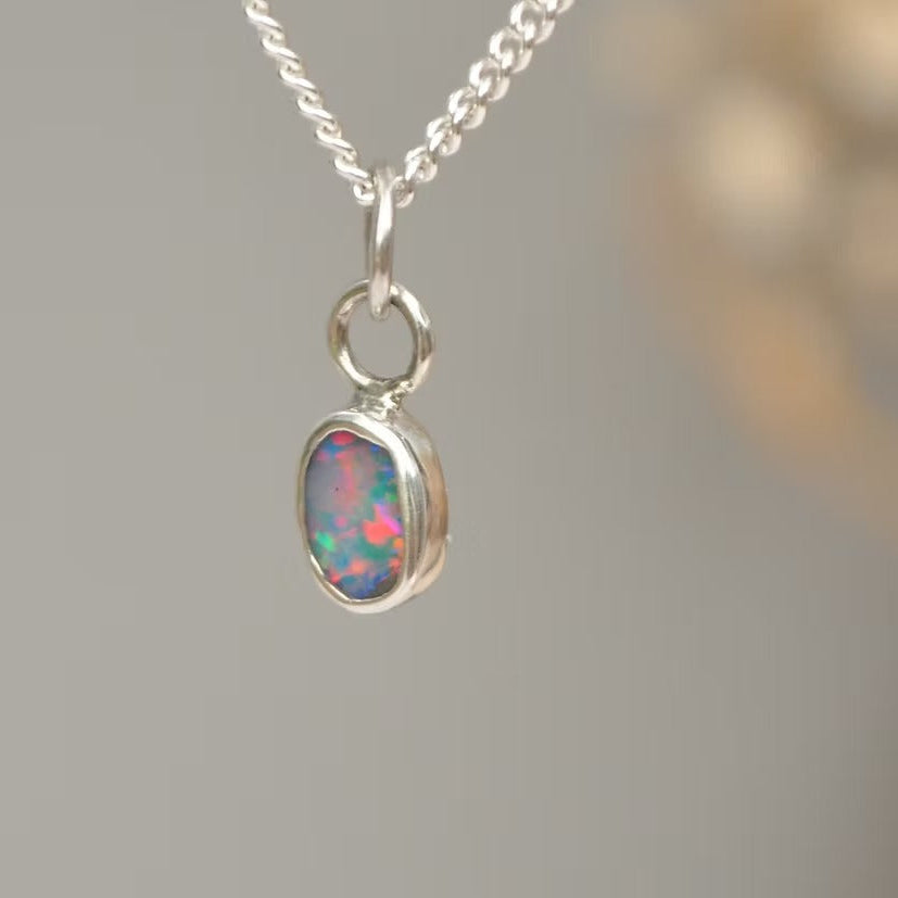 A silver chain necklace with a small oval pendant featuring a colorful opal stone in a silver bezel setting, shown against a soft gray background.
