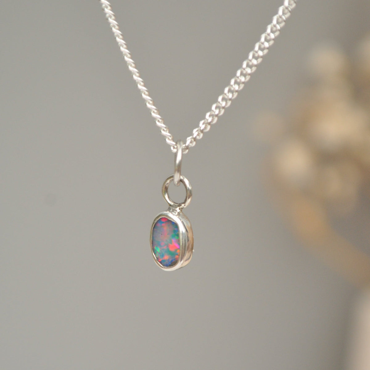 A silver chain necklace with a small oval pendant featuring a colorful opal gemstone, shown against a soft gray background with a blurred decorative element on the right.