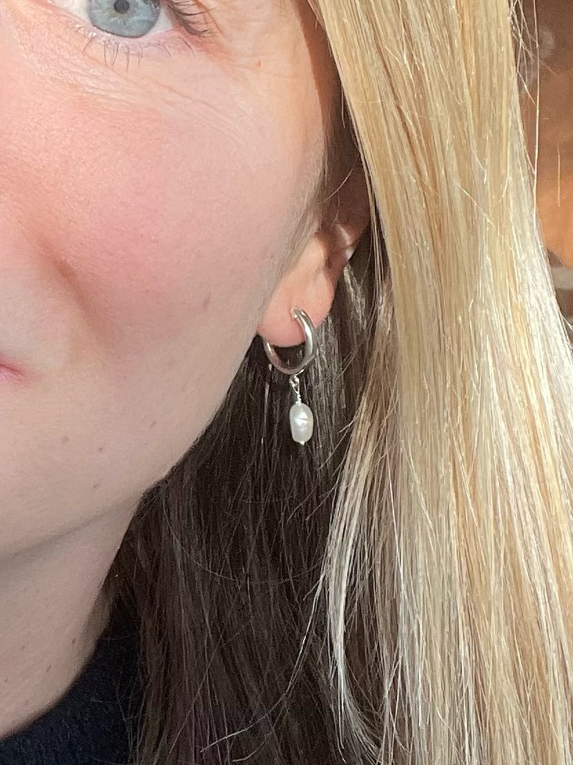 A close-up of a person's ear wearing a silver hoop earring with a dangling white pearl, shown against blonde and brown hair.